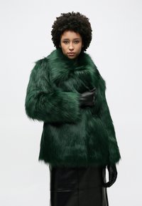 Πράσινο παλτό από ψεύτικη γούνα με ψηλό γιακά, φαρδιά μανίκια και απαλό υφή, συνδυασμένο με μαύρα δερμάτινα γάντια και γυαλιστερή μαύρη φούστα.