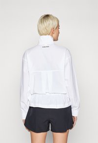Casaco desportivo branco com gola alta, cintura elástica e mangas franzidas; apresenta um costas plissadas e detalhe do logótipo na parte traseira.