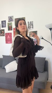 Γυναίκα με μαύρο φόρεμα με βολάν ρυθμίζει το λουράκι μιας κρεμ πλεκτής τσάντας, στέκεται σε ένα διακοσμημένο σαλόνι με γκρι καναπέ και φωτιστικά δαπέδου.