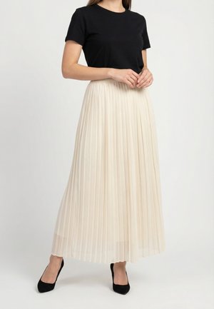 Mujer vestida con un top negro de manga corta, falda larga plisada de color crema y tacones negros de punta puntiaguda, de pie con las manos entrelazadas.