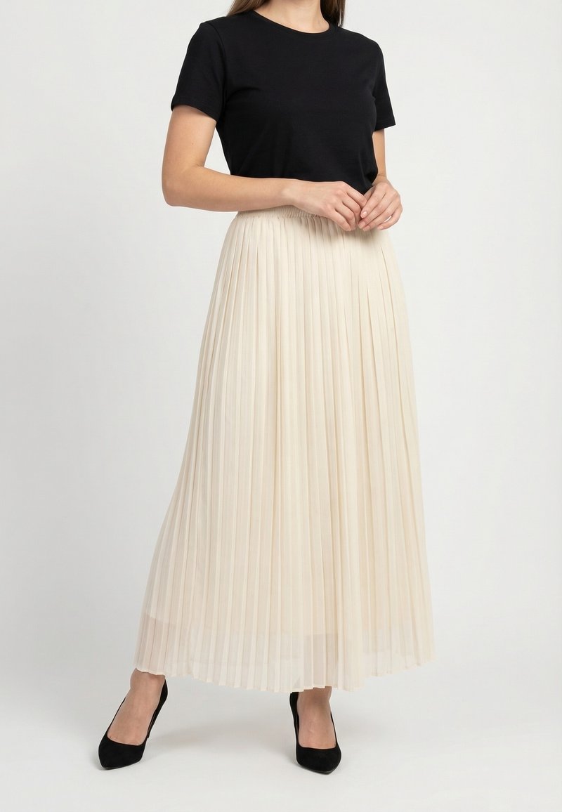 Mujer vestida con un top negro de manga corta, falda larga plisada de color crema y tacones negros de punta puntiaguda, de pie con las manos entrelazadas.