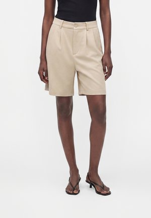 Beige, hoch taillierte, maßgeschneiderte Shorts mit Falten, Vorderknopf und Reißverschluss, kombiniert mit schwarzem ärmellosem Top und schwarzen Absatzsandalen.