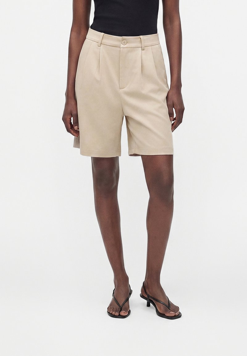 Short beige taille haute cintré avec plis, bouton sur le devant et fermeture éclair, porté avec un haut noir sans manches et des sandales à talons noires.