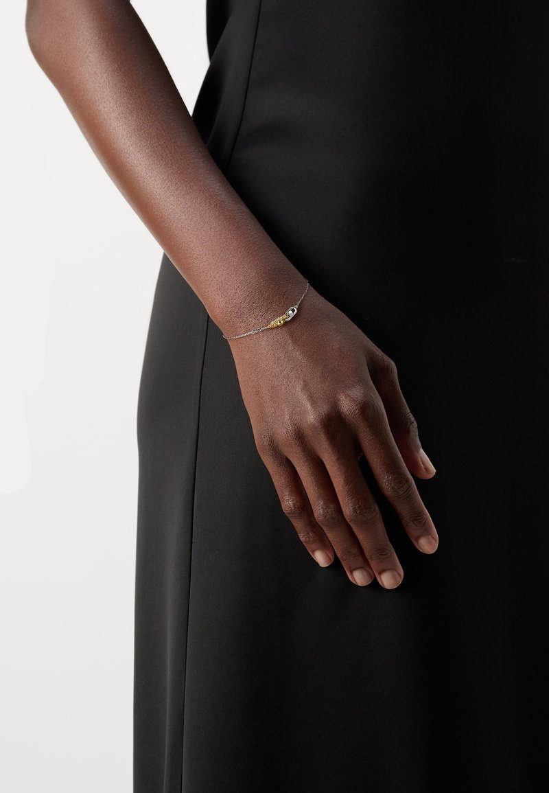 Brazo de piel oscura que lleva una delicada pulsera de plata con cuentas doradas y plateadas apoyada contra una prenda negra.