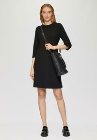 Robe noire longueur genoux avec des manches trois-quarts. Tissu texturé, coupe décontractée. Le mannequin tient un sac noir avec une bandoulière. Elle porte des mocassins noirs.