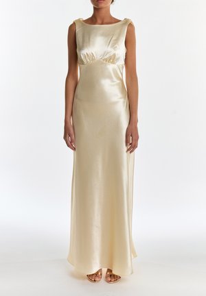 Frau trägt ein bodenlanges, ärmelloses Champagner-Satin-Kleid mit gerafften Details unter der Brust und goldfarbene Sandalen.