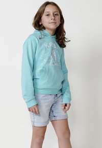 Gaiši zils kapucis ar kangaroo kabatu un baltu grafiku ar haizivi. Saskaņots ar sarullētiem džinsa šortiem. Mīksta, teksturēta auduma.