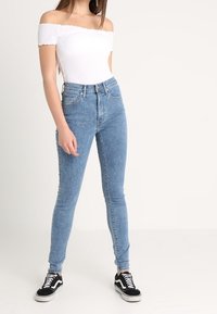 Bodysuit blanco con escote off-the-shoulder combinado con jeans ajustados de cintura alta en un tono azul claro. La modelo lleva zapatillas Vans en blanco y negro.