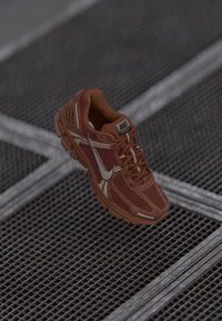 Egyetlen barna Nike futócipő fehér swoosh-szal �és "Vomero 5" felirattal fém rácsos padlón.