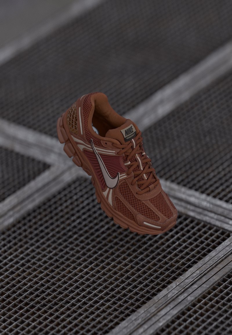 Egyetlen barna Nike futócipő fehér swoosh-szal és "Vomero 5" felirattal fém rácsos padlón.