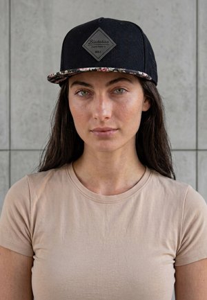 Mladá žena s dlhými tmavými vlasmi, ktorá má na sebe čiernu šiltovku s kvetinovým lemom a béžové tričko, stojaca pred sivou kachličkovou stenou.