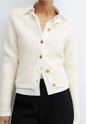 Femme portant un cardigan blanc texturé avec des boutons dorés sur une chemise à col blanc et un pantalon noir.