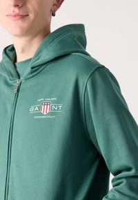 Zeleni zip-up dukserica izrađena od mekanog materijala, s prednjim logom na kojem je dizajn štita. Uključuje veliku kapuljaču i glatku teksturu.