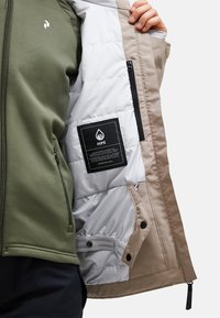 Die Softshell-Jacke verfügt über eine olivgrüne Außenfarbe, ein weißes, isoliertes Innenfutter und beigefarbene Akzente. Sie beinhaltet eine Zipper-Tasche und ein Markenlabel.