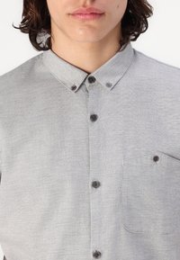 Lichtgrijze overhem shirt met een kraag, een voorzak en donkere knopen, gemaakt van een geweven stof.