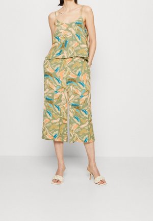 Femme portant un débardeur ample sans manches et un pantalon large court à imprimé floral abstrait vert, associé à des sandales blanches à talons.