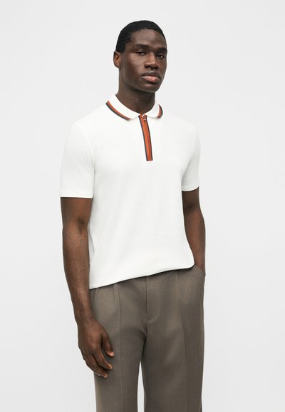 Άνδρας με άσπρο πόλο μπλουζάκι με κοντό μανίκι, με πορτοκαλί και μαύρο φερμουάρ στον γιακά, και καφέ παντελόνι, στέκεται μπροστά σε λευκό φόντο.