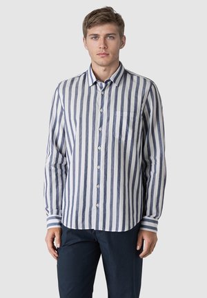 Young man wearing a blue and white vertical striped button-up shirt and dark pants, standing against a plain light background.