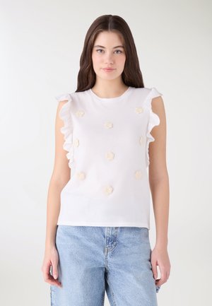 Giovane donna che indossa un top bianco senza maniche con volant ai lati e piccole applicazioni floreali, abbinato a jeans di denim azzurro chiaro.