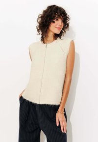 Gilet sans manches de couleur crème en tricot avec une finition texturée, doté d'une fermeture éclair à l'avant, associé à un pantalon à rayures noires.