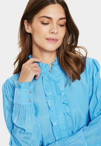 Blusa azzurro chiaro con colletto a volant e apertura frontale. Maniche lunghe a palloncino con dettagli in bottone ai polsini. Tessuto con una superficie liscia.