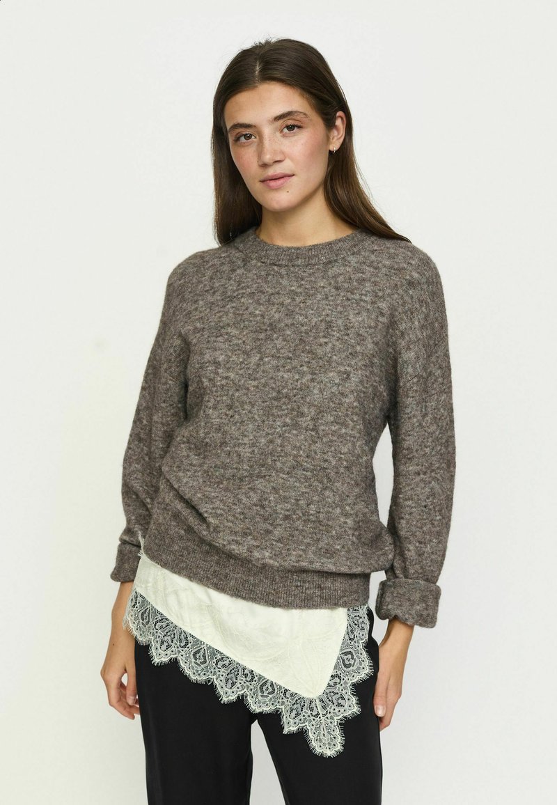 Jeune femme aux longs cheveux bruns portant un pull gris sur un chemisier blanc orné de dentelle et un pantalon noir, debout devant un fond uni.