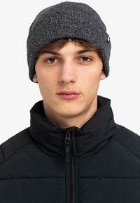 Beanie grigia a maglia con texture a coste, indossato sopra capelli scuri, abbinato a una giacca nera con zip e colletto alto.
