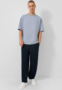 Man in een lichtblauwe oversized t-shirt, donkerblauwe wijde broek en witte sneakers, staand tegen een effen lichtgrijze muur.