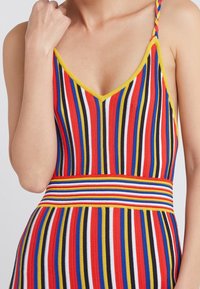 Femme portant une robe sans manches à rayures multicolores avec des bretelles tressées et une taille ajustée, main près de la clavicule.