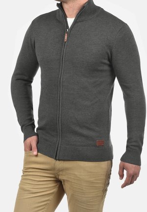 Mężczyzna ubrany w ciemnoszary sweter na zamek z prążkowanymi mankietami i dołem, w beżowych spodniach, z ręką w kieszeni na białym tle.
