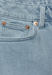 Jeans en denim bleu clair avec une texture lisse, présentant deux rivets de couleur cuivre et une ceinture cousue.