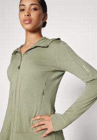 Sweatshirt com fecho em verde oliva, com um design ajustado, tecido texturizado, bolsos laterais e um capuz desportivo. Inclui uma marcação discreta na manga.