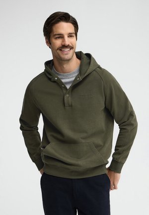 Hombre sonriente con cabello oscuro y bigote que lleva una sudadera con capucha verde oliva sobre una camiseta gris y pantalones oscuros, de pie con las manos en los bolsillos.
