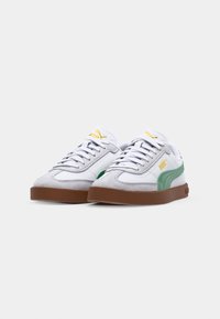 Sneaker bianche con accenti in suede grigio, striscia laterale verde e suola in gomma marrone. Presentano lacci bianchi e un logo giallo sulla linguetta.
