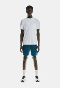 White t-shirt with a small logo on the left chest, paired with blue shorts. The model wears white socks and black and white running shoes.