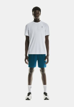 Vit t-shirt med en liten logotyp på vänster bröst, tillsammans med blå shorts. Modellen har på sig vita strumpor och svarta och vita löparskor.