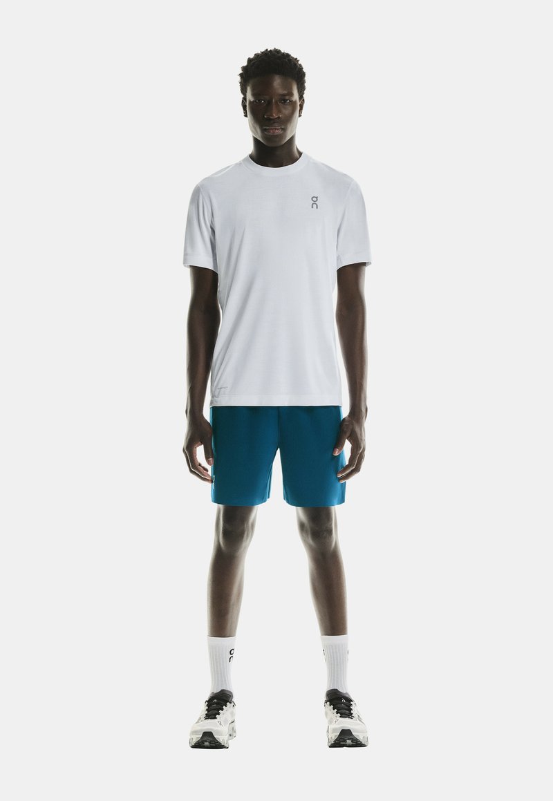 White t-shirt with a small logo on the left chest, paired with blue shorts. The model wears white socks and black and white running shoes.
