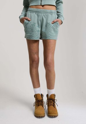 Person wearing light teal floral-patterned shorts, matching cropped sweatshirt, white socks, and tan lace-up boots. Hands in pockets.