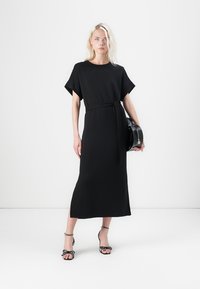 Černé, středně dlouhé šaty s krátkými rukávy, úzkým pasem a hladkým materiálem. Modelka má na sobě černé lodičky s ozdobnými pásky.