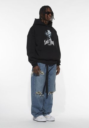 Black hooded sweatshirt with central graphic text "Lost Youth," paired with distressed light blue jeans and white sneakers. Sunglasses included.