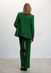 Femme aux cheveux mi-longs portant un costume vert sur mesure et des sandales à talons noires, debout devant un fond clair uni.