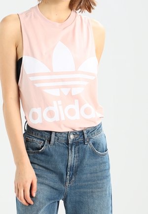 Kobieta w różowej bezrękawniku Adidas z białym logo i niebieskich dżinsach z wysokim stanem, stojąca na tle jasnego, jednolitego tła.