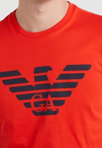 Camiseta de algodón roja con un gran gráfico negro que presenta un emblema de águila y el texto "GA", sobre un fondo liso y plano.
