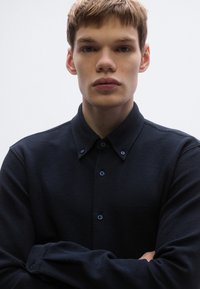 Tengerészkék, texturált ing galléros nyakkivágással, elöl gombokkal és finom márkajelzéssel. Az anyag könnyűnek és légáteresztőnek tűnik.