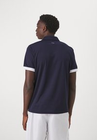 Mørkeblå polo skjorte med hvite detaljer ved kragen og ermene. Har en glatt tekstur og FILA-logoen på baksiden av halsåpningen.