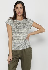 Una camiseta de manga corta, gris con un patrón, presenta un diseño geométrico con acentos de texto en negro. Tela suave con un corte relajado, combinada con pantalones verde oliva.