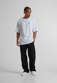 Hvid bomulds t-shirt med korte ærmer, der har et minimalistisk sort logo. Sammen med sorte bukser og hvide sneakers. Ensfarvet baggrund.
