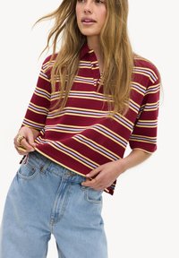 Femme portant un polo rayé bordeaux et un jean taille haute bleu clair, ajustant le bas de son polo avec ses mains.