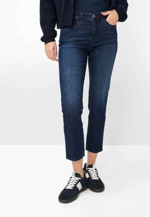 Person iført mørkeblå cropped jeans, sort-hvide sneakers og en mørk langærmet top, stående med den ene hånd i lommen.