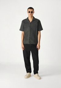 Svart skjorte med korte ermer, krage og knapper foran, kombinert med svarte bukser og lyse joggesko; modellen har på seg mørke solbriller.